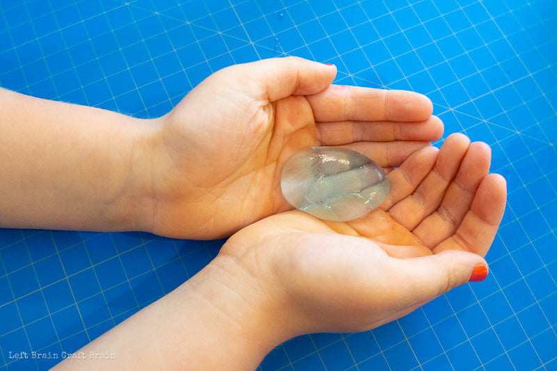 Small, white, female hands cradle an edible water bubble while resting on a blue counter with white graph lines. Credit to Left Brain Craft Brain